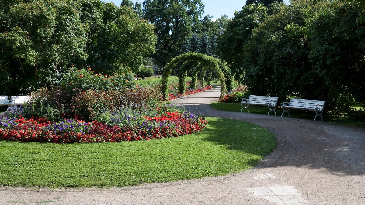 En stor runt blomsterodling med en grusgång runt. Vid sidan finns bänkar, träd och buskar.