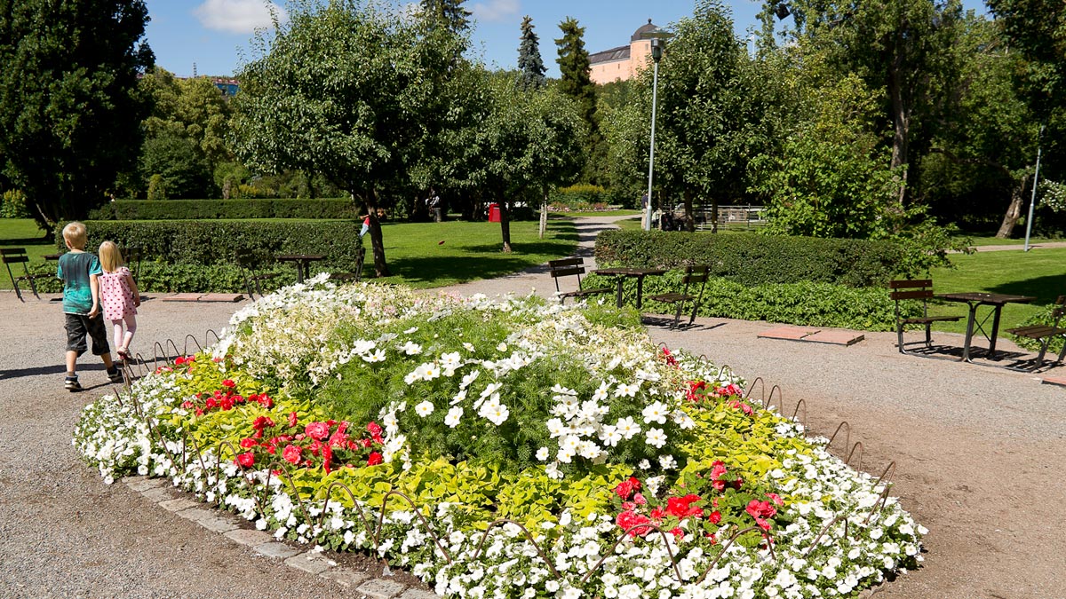 Två barn går på en grusgång mellan cafébord och blomsterodlingar. I bakgrunden syns träd, buskar och Uppsala slott.
