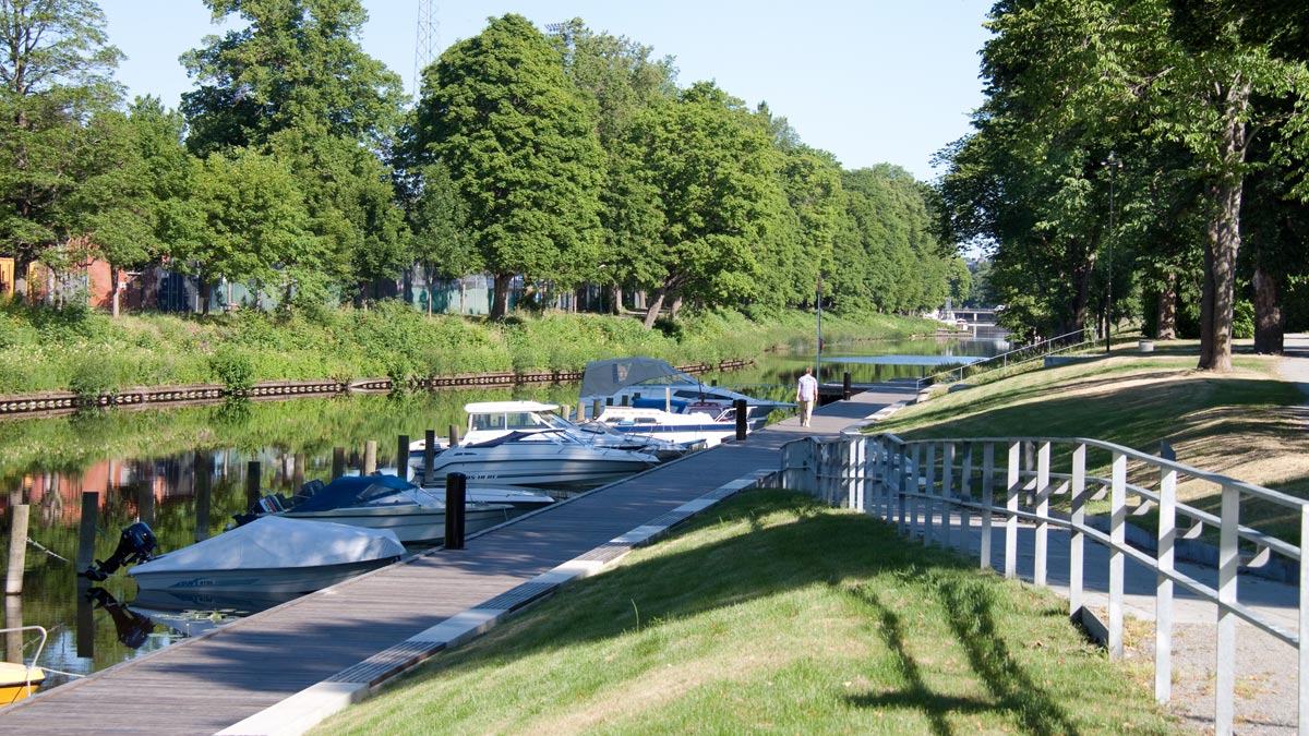 Några båtar förtöjda vid en träbrygga längs med Fyrisån. Längs med ån finns stora lövträd.