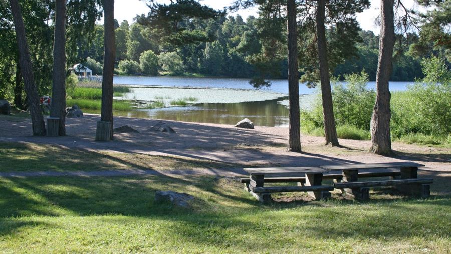 Två bänkar och några träd på gräsytan vid Sunnerstabadets sandstrand.