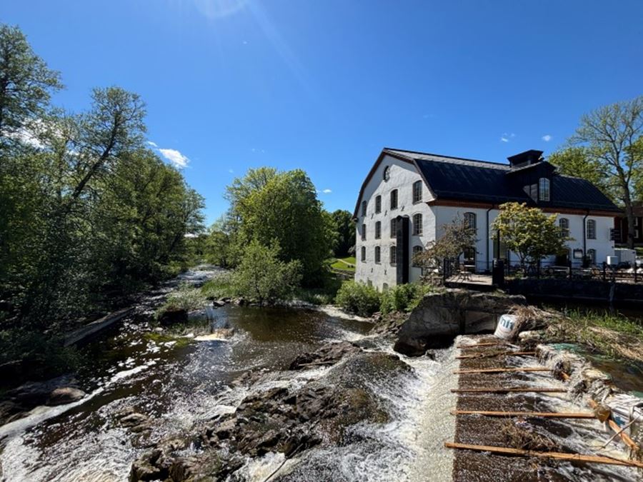 Vitt brukshus vid en porlande å med liten fors i förgrunden, omgivet av gröna träd under klarblå himmel.