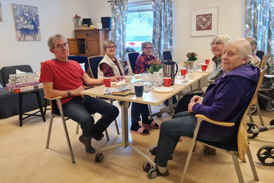 En grupp personer sitter runt ett bord och fikar och samtalar i en hemtrevlig samlingslokal.