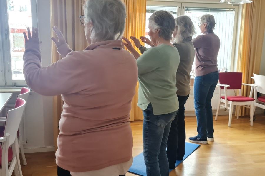 Fyra personer står på rad i ett rum och gör lugna rörelseövningar framför ett fönster.