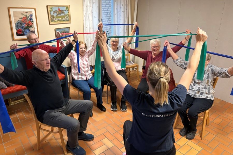 En grupp personer sitter i en cirkel och tränar med färgade träningsband under ledning av en instruktör.