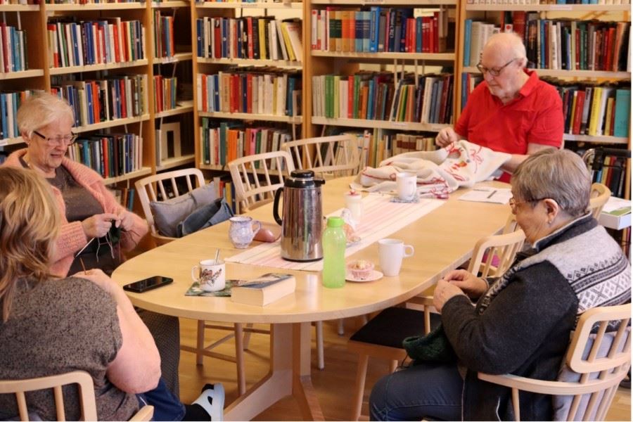 En grupp personer sitter runt ett bord i ett bibliotek och fikar medan några handarbetar.