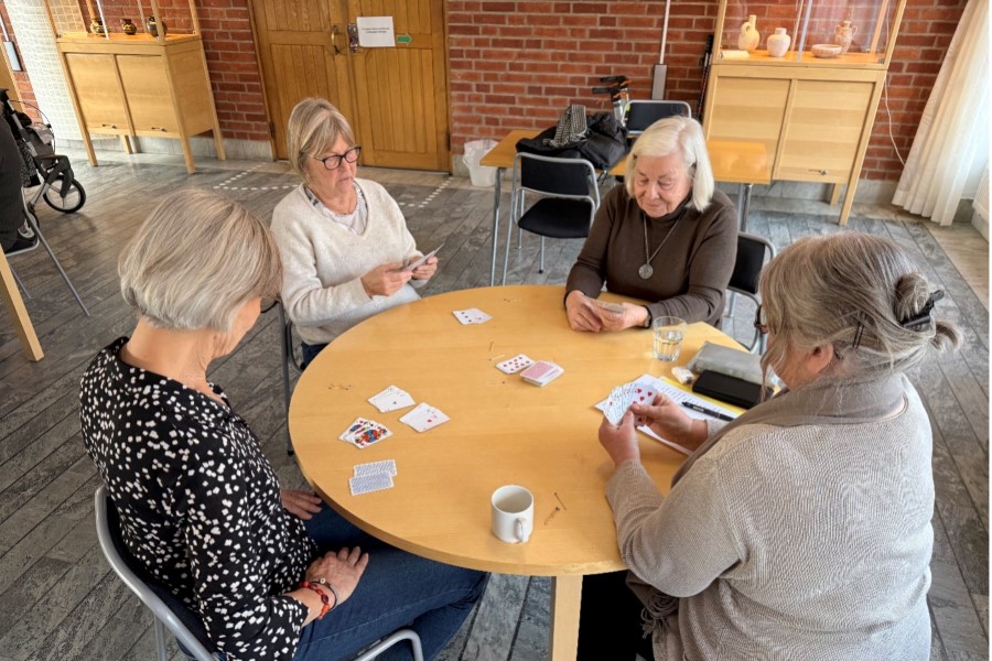 Fyra personer sitter runt ett bord och spelar kort i en ljus lokal.