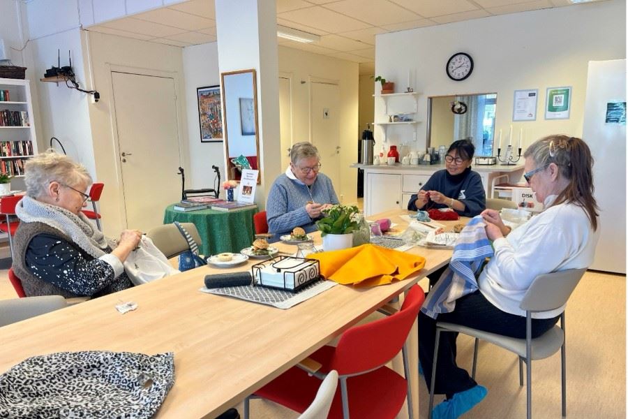 En grupp personer sitter runt ett bord och handarbetar i en hemtrevlig lokal.