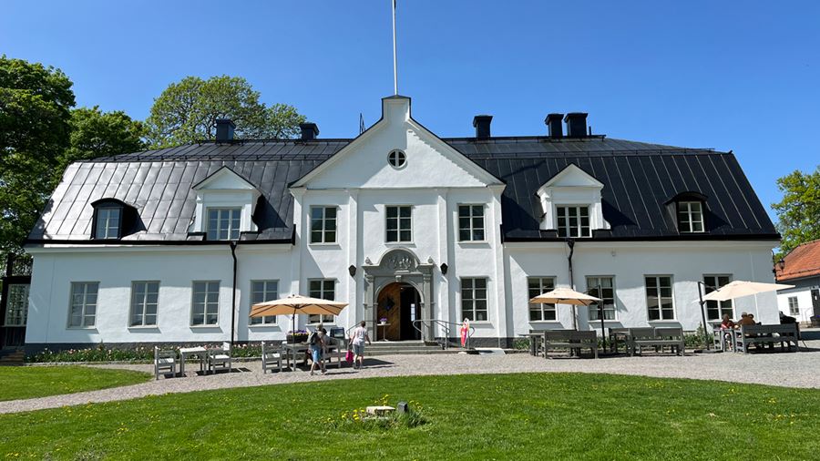 Herrgårdsbyggnaden i Hammarskog. Det är en stor vit byggnad med putsad fasad och svart plåttak. Herrgårdens café har sittplatser med parasoll utanför byggnaden.