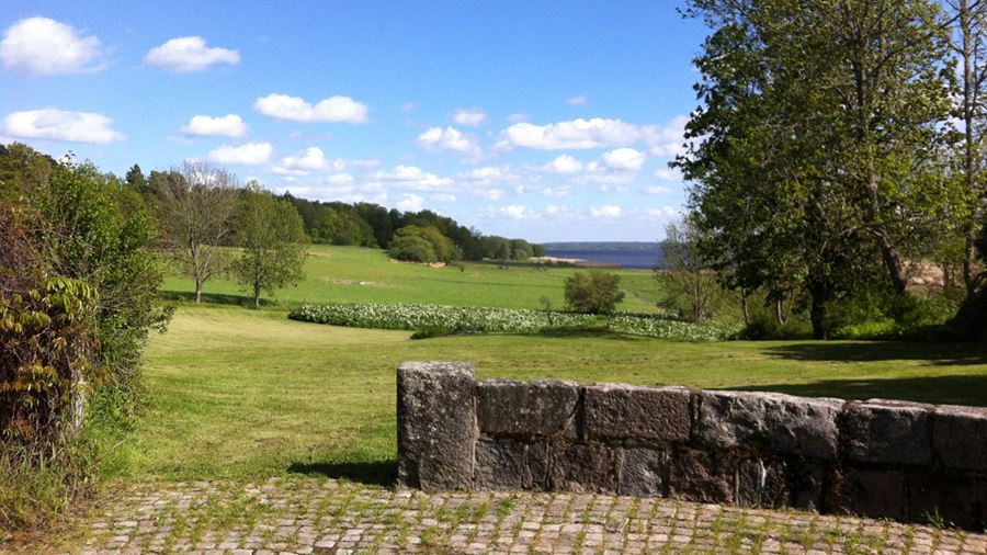 I förgrunden en låg gammal stenmur. Bakom den sträcker sig stora gräsytor ned mot vattnet i Hammarskogs friluftsområde.