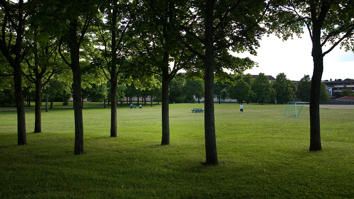En stor grästyta med träd, ett fotbollsmål och en bänk. Tre personer syns längre bort.